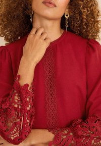 Frau mit lockigem Haar, die goldene Ohrringe und eine rote Bluse mit filigranen Spitzensäumen an den Ärmelenden und einem Spitzenbesatz vorne trägt.