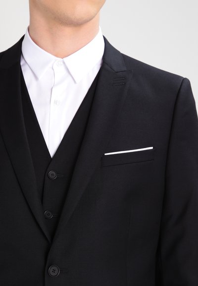 Zwarte pak met een gestructureerde afwerking, scherpe revers en een wit overhemd eronder. Beschikt over een vest met drie knopen en een zakdoekzakje met een witte accent.