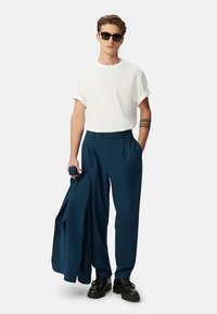 Witte T-shirt met opgerolde mouwen, marineblauwe geplooide broek, een donker colbert vasthoudend, en voorzien van zwarte zonnebril en schoenen. Soepele stof, op maat gemaakte pasvorm.