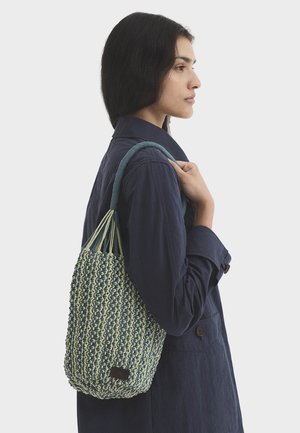 Vrouw met een marineblauwe jas draagt een blauw-beige geweven schoudertas, staand in profiel tegen een witte achtergrond.