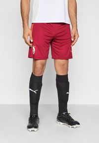 Weinrote Fußballshorts mit Logo und Sternen, kombiniert mit schwarzen knielangen Socken und schwarzen Stollenschuhen mit strukturierter Oberfläche.
