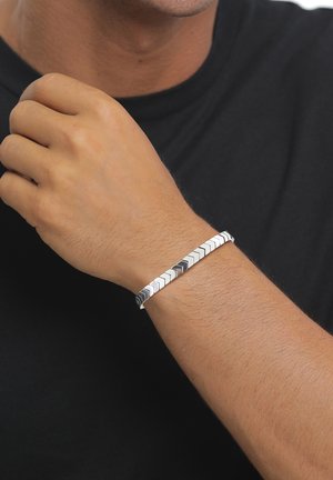 Wrist with a silver bracelet of interlocked chevron-shaped links worn on a light brown arm against a black shirt background.