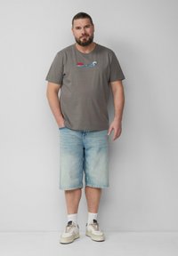 Grijze katoenen T-shirt met een "JAWS" golfafbeelding, gecombineerd met lichtblauwe denim shorts en witte sneakers met beige accenten.