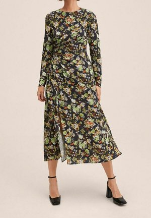 Robe à manches longues à motifs floraux sur une base noire, avec des motifs vibrants de vert, orange et blanc, fendue sur un côté, accompagnée de chaussures à talons noires.