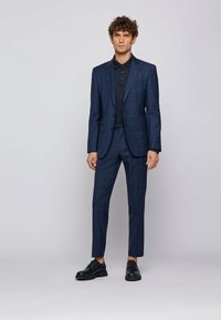 Navy checkered suit with a tailored jacket and slim fit trousers, paired with a black polo shirt and black shoes, on a neutral background.