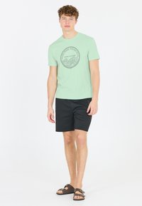 Ljusgrön bomullströja med en rund grafisk design som visar vågor och text. Svarta shorts och bruna sandaler kompletterar outfiten.