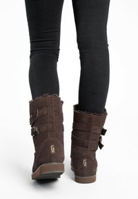Botas de ante marrón hasta el tobillo con forro de piel, hebillas laterales y suela de goma plana, combinadas con jeans ajustados negros sobre un fondo blanco.