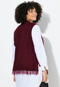 Chaleco de punto burdeos con dobladillo con flecos, llevado sobre una camisa blanca con puños abotonados. Presenta un cuello alto y un ajuste regular.