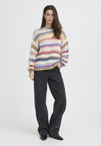 Pull en maille avec rayures multicolores dans des tons pastels, coupe ample, poignets côtelés, associé à un jean noir à jambes larges et des chaussures noires pointues.