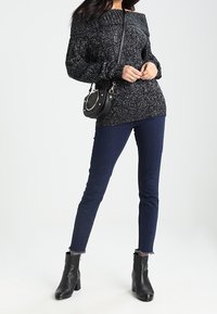 Suéter de punto en negro y gris moteado con diseño de hombros descubiertos, combinado con jeans skinny azul oscuro y botines negros. Acentuado con un pequeño bolso negro.
