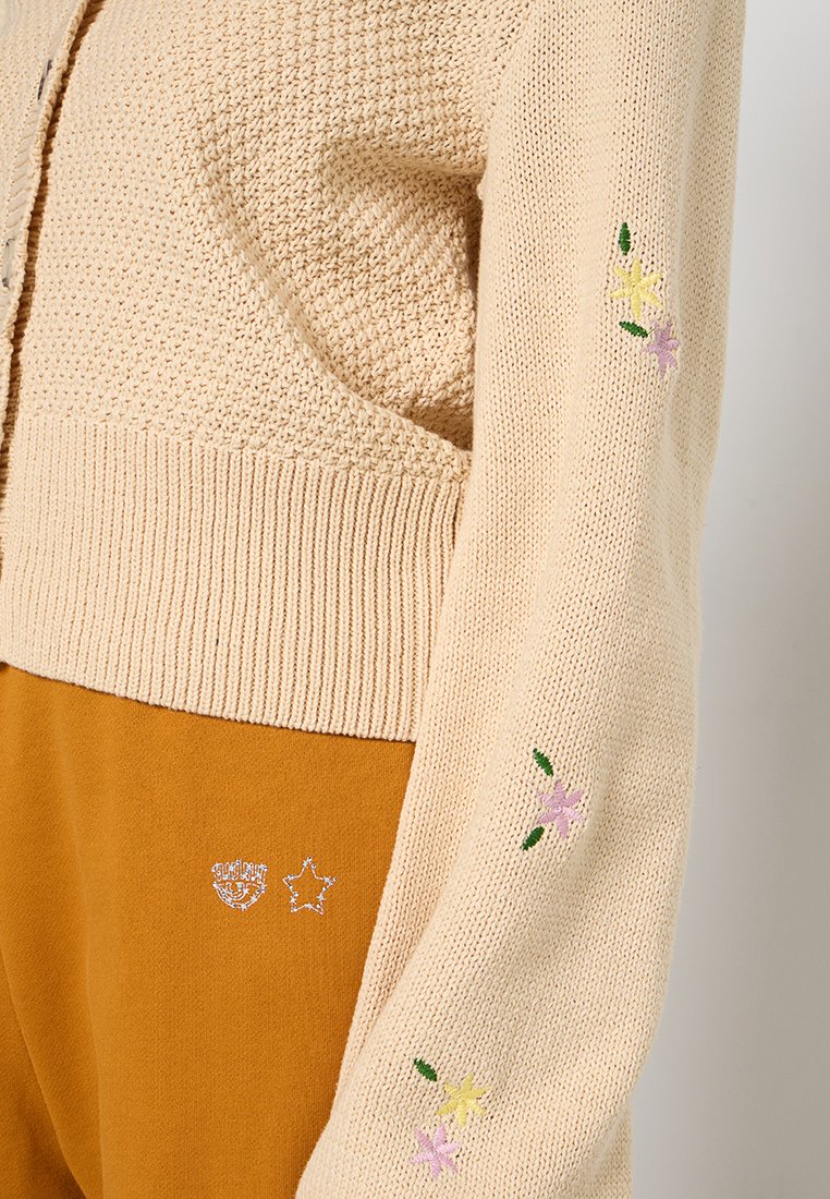Beige gebreide cardigan met een textuurafwerking, voorzien van bloemstickers in pastelkleuren op de mouwen en een geribbelde zoom.