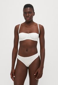Balta bikini komplekts, kas ietver strukturētu bandeau topu ar stiepjamiem elementiem un atbilstošām zemu augšpusi biksēm. Gluda auduma, minimāla dizaina izstrādājums.