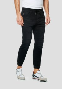 Czarne spodnie dresowe o zwężanym kroju, z elastycznym paskiem w talii i bocznymi kieszeniami. Na kolanach ozdobione przeszyciami. Noszone z białymi i szarymi trampkami.