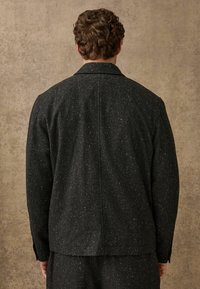 Jachetă texturată neagră cu un model discret punctat, cu o croială ajustată și o deschidere unică la spate. Fără închideri vizibile.