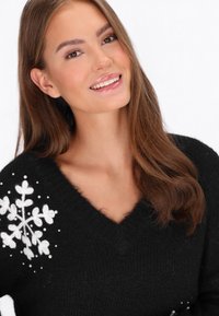 Schwarzer gestrickter Pullover mit V-Ausschnitt, der weiße Blumenstickerei und dekorative Perlen auf der linken Schulter hat. Weicher Stoff.