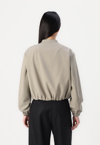 Giacca cropped beige chiaro con colletto alto, vita elasticizzata e maniche lunghe. Il tessuto ha una finitura liscia e opaca.