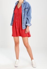 Röd klänning med prickar och åtsittande midja, lagd under en ljusblå denimjacka. Outfiten kompletteras med vita sneakers för en avslappnad look.