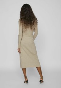 Robe beige tricotée avec des manches longues, taille cintrée et ourlet mi-long. Présente une texture côtelée et une finition lisse, portée avec des talons ouverts.