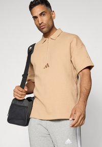 Beige textuur polo shirt met een half-rits, voorzien van een centraal logo detail. Model houdt een kleine zwarte schoudertas vast. Grijze joggingsbroek.
