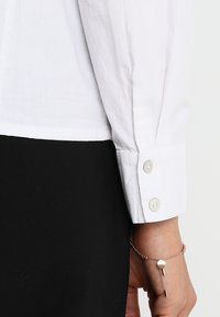 Witte button-up shirt met lange mouwen, voorzien van ronde knopen bij de manchetten. Het shirt is iets doorzichtig met een gladde textuur en gecombineerd met zwarte stof.