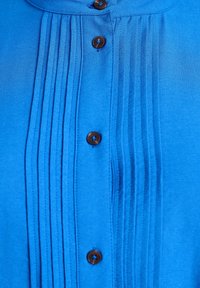 Sinine särk, millel on volditud esiosa, pruunid nööbid ja pehme tekstuur. Krae on ümar ja kangas tundub sile ja kerge.