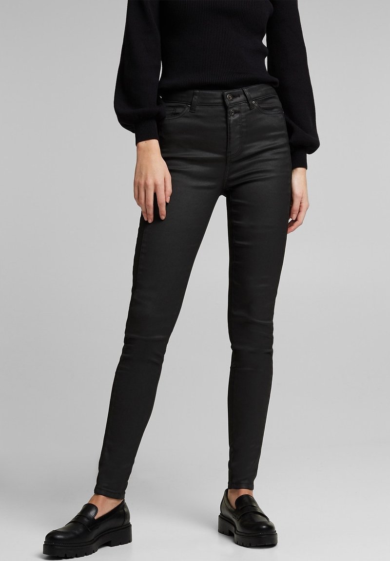 Pantalon noir ajusté taille haute associé à des mocassins noirs et un haut manches longues noir sur une personne debout.