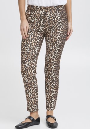 Vrouw die een slim-fit broek met luipaardprint en zwarte Mary Jane schoenen met gespen draagt, staand tegen een effen lichte achtergrond.
