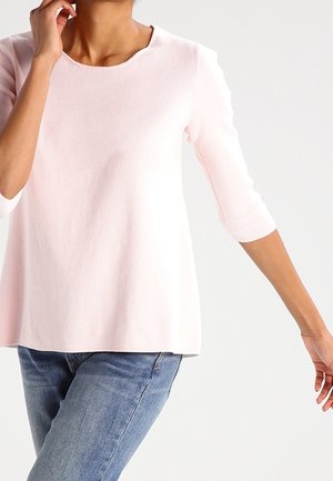 Osoba ubrana w jasnoróżową koszulę z długim rękawem i niebieskie jeansy, jedna ręka przy twarzy, a druga opuszczona w dół.