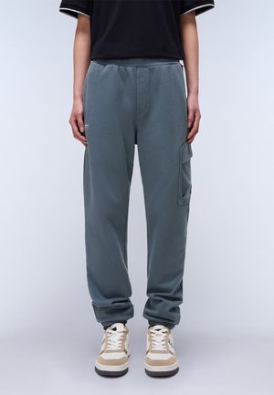 Persoon draagt donkergrijze cargobroek met elastische boorden en beige-witte sneakers, staand tegen een effen lichte achtergrond.