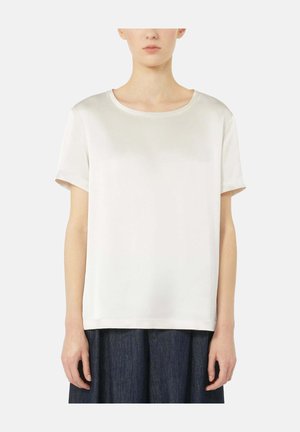 Vrouw die een effen wit T-shirt met korte mouwen en ronde hals draagt en een donkere geplooide rok, staand tegen een witte achtergrond.