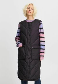 Vadderad svart ärmlös väst med diagonala sömmar, med framfickor och knappstängningar, bärs över en flerfärgad randig tröja.