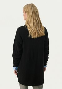 Pulover oversized negru, cu mâneci lungi și guler rotund. Prezintă manșete și tiv cu nervuri, asociat cu o cămașă bleu deschis dedesubt.
