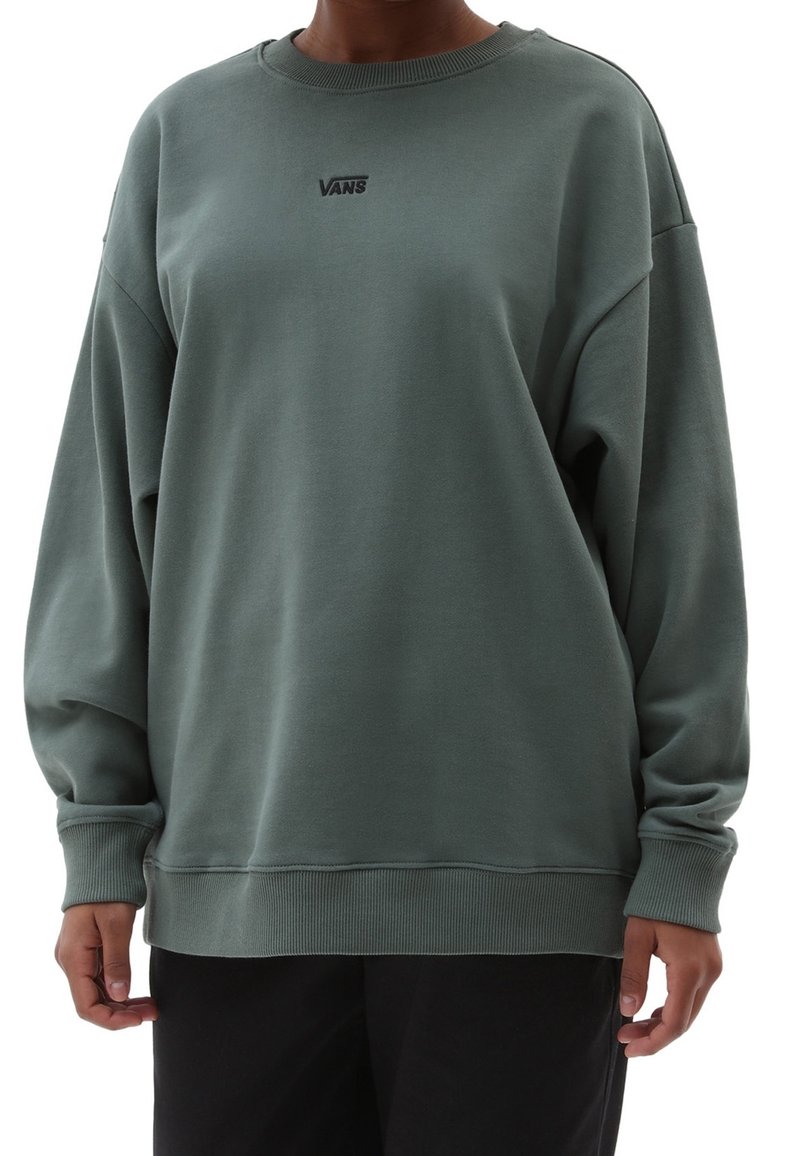 Personne portant un sweat-shirt Vans vert foncé oversize et un pantalon noir, debout avec les bras détendus le long du corps sur fond blanc.