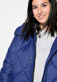 Chaqueta acolchada azul con acabado texturizado, con cierre de cremallera, botones a presión y capucha desmontable. Usada sobre una camiseta gris claro.