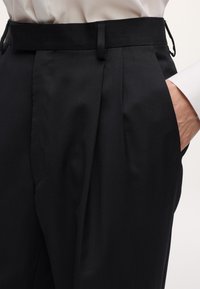 Pantaloni sartoriali neri con tasche laterali indossati con una camicia bianca, mano parzialmente in tasca, che mostra le passanti per la cintura e le pieghe frontali.