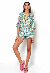 Kurzärmeliges, knöpfbares Hemd mit einem bunten wellenförmigen Muster in Grün, Pink und Orange, kombiniert mit hellen Denimshorts und leuchtend pinken Sandalen.