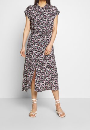 Frau trägt ein schwarzes, florales Midikleid mit kurzen Ärmeln, durchgehender Knopfleiste, Taillengürtel und weißen Riemchensandalen, steht vor einem einfarbigen Hintergrund.