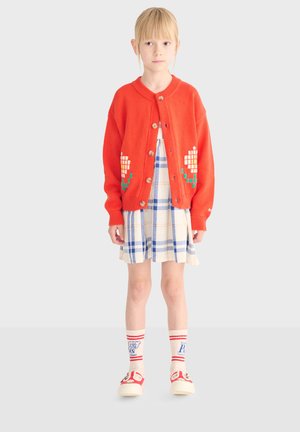 Mladé dievča v oranžovom kardigane s bielymi a zelenými pixelovanými kvetmi, v modro-bielych kockovaných šatách, bielych ponožkách a červených teniskách.