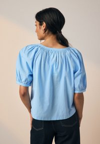 Hellblaues Kurzarmoberteil mit Puffärmeln und gerafftem Ausschnitt. Hergestellt aus strukturiertem Stoff mit dezenten Mustern.