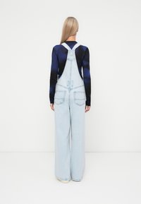 Lichtblauwe denim overall met brede benen, verstelbare bandjes en twee achterzakken. Draag over een donker, lang mouwtop met een blauw patroon.