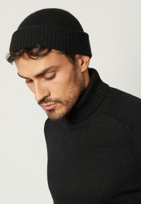 Svart ribbad stickad mössa med uppvikt kant, parat med en svart tröja med hög hals. Båda plagg har en slät textur och minimalistisk design.