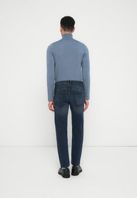 Hombre vistiendo un suéter de cuello alto azul, jeans azul oscuro y zapatos negros, de pie frente a una pared blanca con las manos relajadas a los lados.