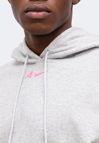 Κοντινό πλάνο ενός ατόμου που φοράει ανοιχτό γκρι φούτερ με λευκά κορδόνια και μικρό ροζ λογότυπο Nike στο στήθος.