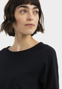 Zwarte lange mouw top met een ronde halslijn, met een gladde textuur en raglan naden. Model kijkt opzij tegen een effen achtergrond.