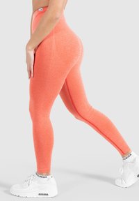 Hoge taille, aansluitende leggings van feloranje stof met een gestructureerd patroon en contrasterende naaddetails, gecombineerd met witte sneakers.