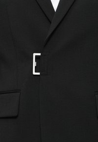 Μαύρο σακάκι με ραφές, με ασημένιο ορθογώνιο κούμπωμα μπροστά και τσέπες με καπάκια και στις δύο πλευρές.