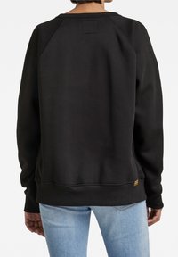 Sweatshirt nero con maniche a raglan, orlo e polsini a coste. Presenta un piccolo dettaglio a patch sulla parte superiore della schiena e un tessuto morbido e texturizzato.