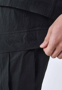 Sort tekstureret stof med plisseret design; har et præget "Project x Paris" logo på en rem. Nærbillede af en hånd, der holder beklædningen.