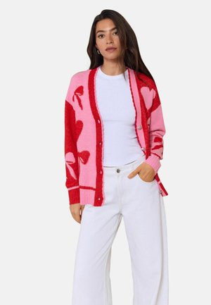 Frau trägt eine pinke und rote Strickjacke mit Herz- und Schleifenmustern, weißes T-Shirt und weiße Hose, steht mit einer Hand in der Tasche.