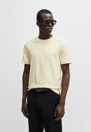 Cremfarbener T-Shirt mit rundem Ausschnitt und kurzen Ärmeln, kombiniert mit schwarzen Hosen und dunkler Sonnenbrille.